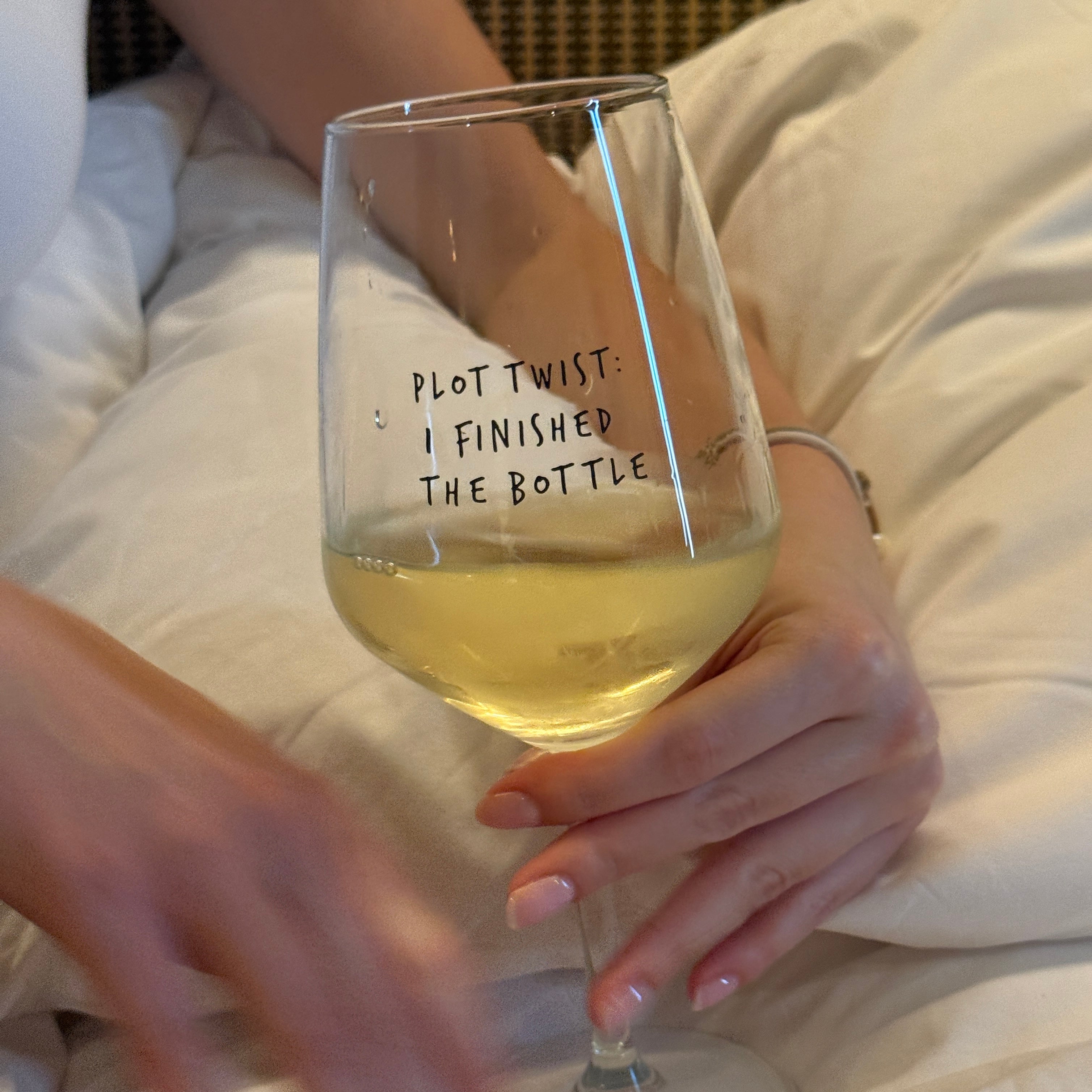 Hand holding the Plot Twist glass filled with white wine in bed