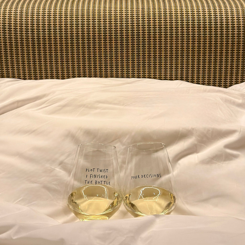 Two wine glasses - Pour Decisions and Plot Twist filled with white wine on a striped bed