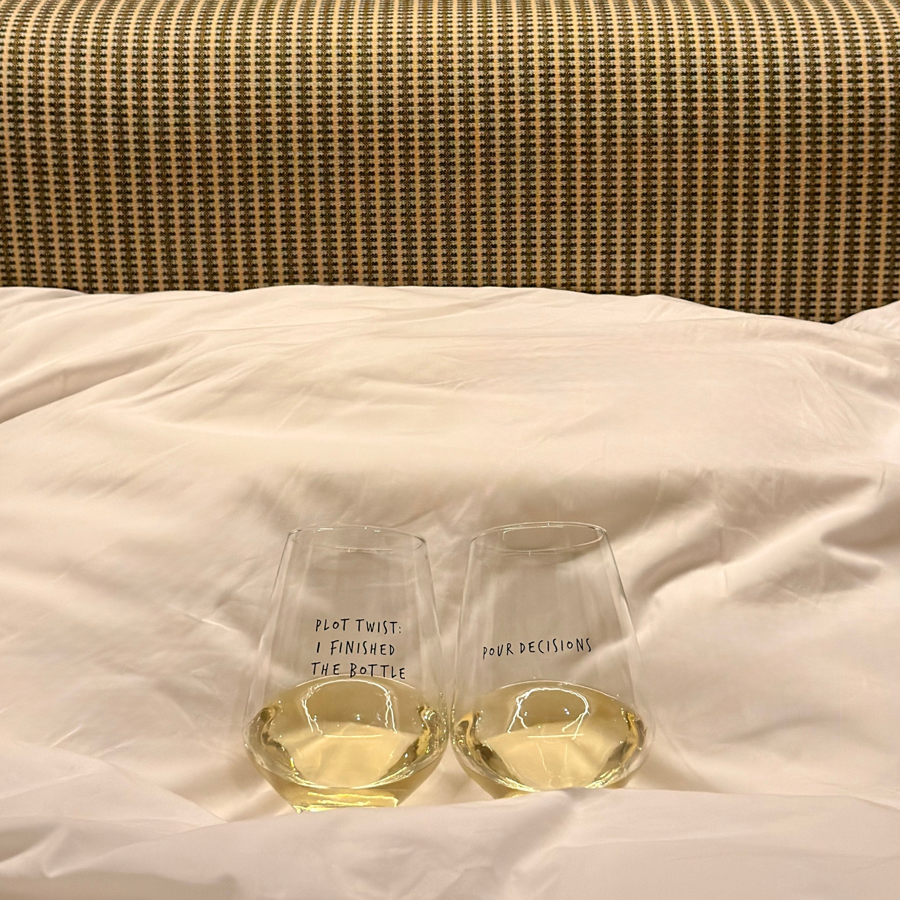 Two wine glasses - Pour Decisions and Plot Twist filled with white wine on a striped bed